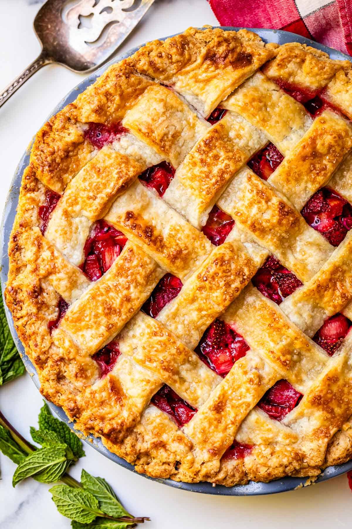 vertical shot of a whole, baked, lattice crust strawberry rhubarb pie with a serving pie serving spoon.