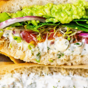 Close-up of a veggie sandwich stacked with spinach, sprouts, tomato, red onion, avocado spread, and creamy sauce on crusty bread. A bold yellow banner at the top highlights VEGGIE SANDWICH.