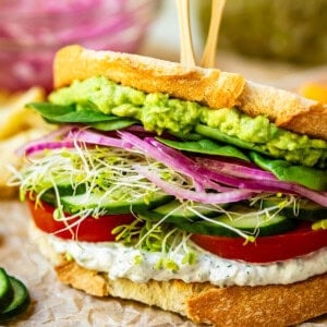 a delicious fresh veggie sandwich with 2 bamboo sandwich skewers in the top on parchment paper.