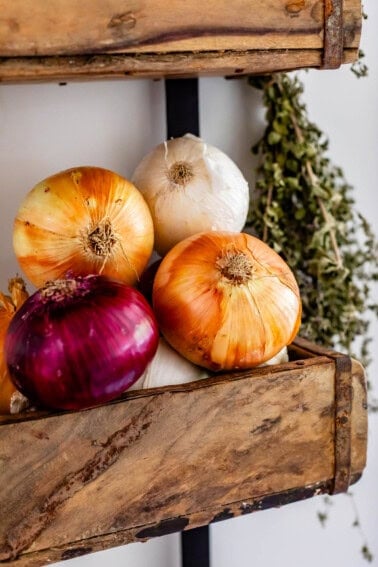 red, white, and yellow onions stacked in a basket on the wall with dried thyme hanging next to it.