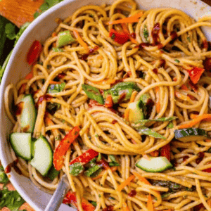 A bowl of sesame noodles with sliced cucumbers, red bell peppers, and carrots, tossed in a savory sauce, with a fork resting inside. Text over the image reads Sesame Noodles.
