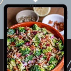 A vibrant broccoli salad with bacon, red onions, and sunflower seeds sits in an orange bowl, displayed on a tablet screen. The text above reads “Healthy Broccoli Salad.”.