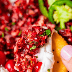 A close-up of a cracker topped with creamy cranberry jalapeno dip and a chunky cranberry-jalapeno mixture, with sliced jalapenos and more dip in the background. Text at the top reads Cranberry Jalapeno Dip.