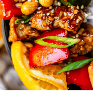 Close-up of a bowl of Kung Pao chicken with chicken, red bell pepper, zucchini, peanuts, green onions, sesame seeds, and orange slices. Text below reads: Easy Homemade Kung Pao Chicken - The Food Charlatan, featuring vibrant Asian flavors.