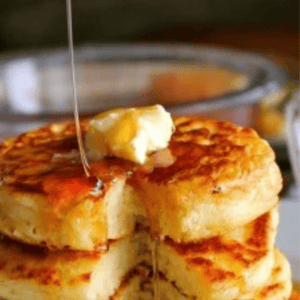 A stack of thick, fluffy old fashioned pancakes topped with butter and syrup being poured over them, with a blurred bowl in the background. Text above reads Old Fashioned Pancakes - The Food Charlatan.