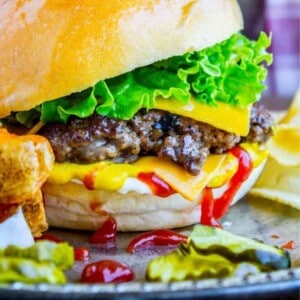 A close-up of a smash burger with melted cheese, lettuce, and pickles on a bun, served with fries and ketchup. A drink sits nearby on a checkered background. The text reads Homemade Smash Burgers.