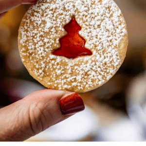 Linzer cookie held up with two fingers in front of christmas lights.