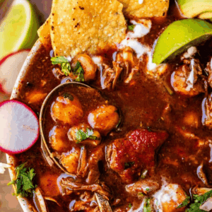 Close-up of a bowl of pozole with shredded meat, hominy, sliced radishes, avocado, tortilla chips, cilantro, and a spoon, topped with a drizzle of cream. Text above reads “Classic Pozole.”.