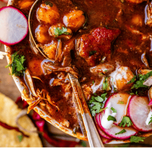 overhead picture of posole rojo in a white bowl with tostada and spoon and limes.
