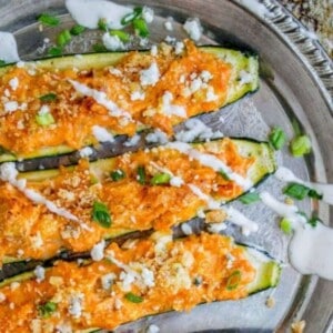 A silver tray holds six zucchini boats filled with a spicy buffalo chicken mixture, topped with crumbled cheese, green onions, and a drizzle of creamy white sauce. The text reads Buffalo Chicken Zucchini Boats—The Food Charlatan.