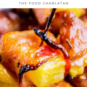 Close-up of Bacon Wrapped Pineapple pieces with crispy bacon around bright yellow pineapple chunks, caramelized edges, and a glossy finish. Text above reads, “Bacon Wrapped Pineapple - The Food Charlatan.”.