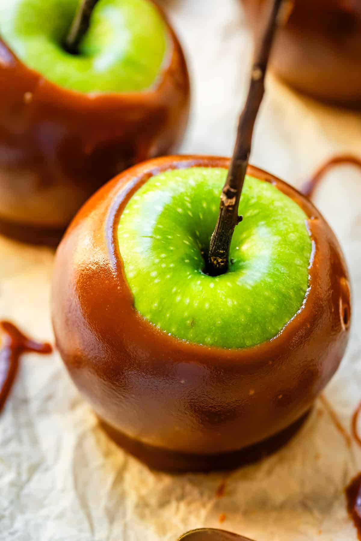 Green caramel apple with a stick in the center on parchment paper.