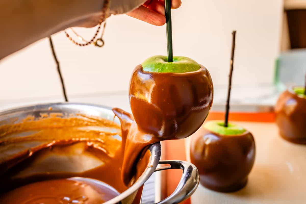 hand holding an apple by the stick, scraping the excess caramel off the bottom on side of pan.