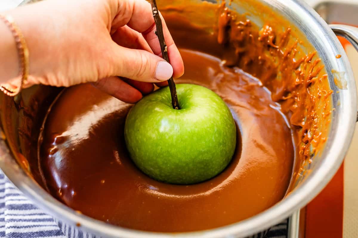 hand lowering an apple by it's stick into a large pot of hot caramel.