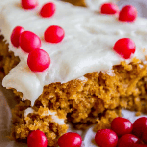 A close-up of pumpkin bars topped with white frosting and red candies, with Pumpkin Bars, Pumpkin cinnamon rolls, and The Food Charlatan at the bottom of the image.