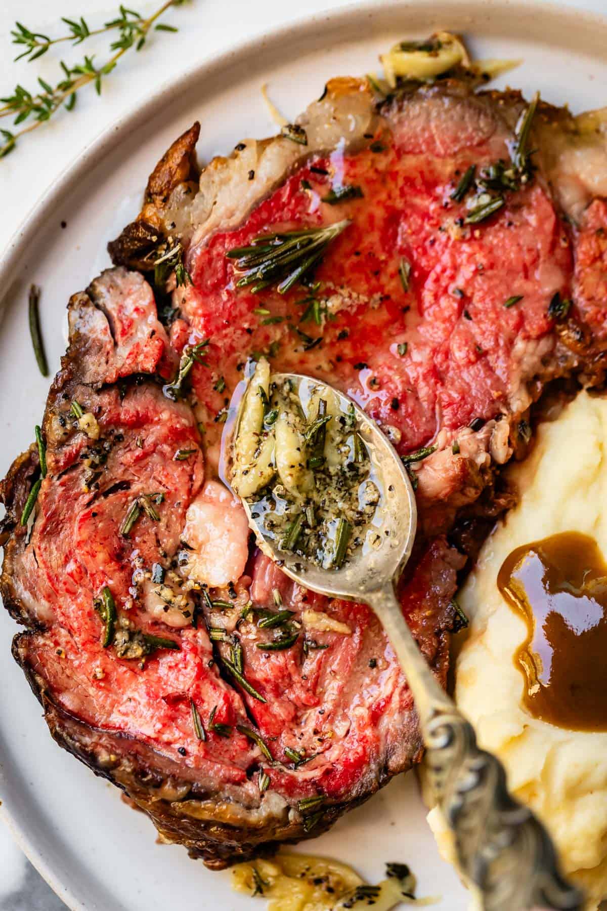 Spooning garlic herb butter onto slice of prime rib on white plate next to mashed potatoes and gravy.