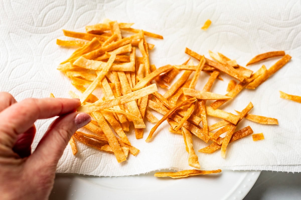 fingers holding a fried tortilla strip, with a pile of fried strips on a paper towel behind it.
