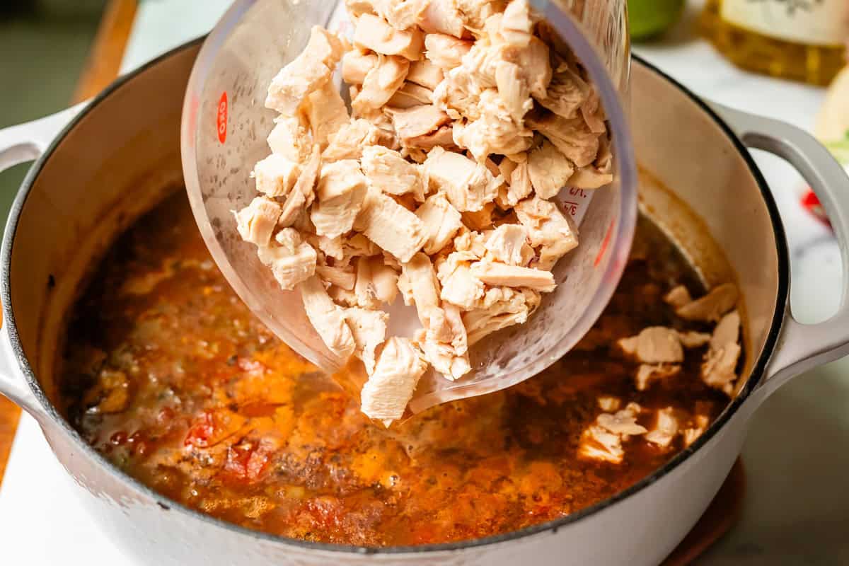 a large measuring cup of chopped chicken being poured into the pot of soup.