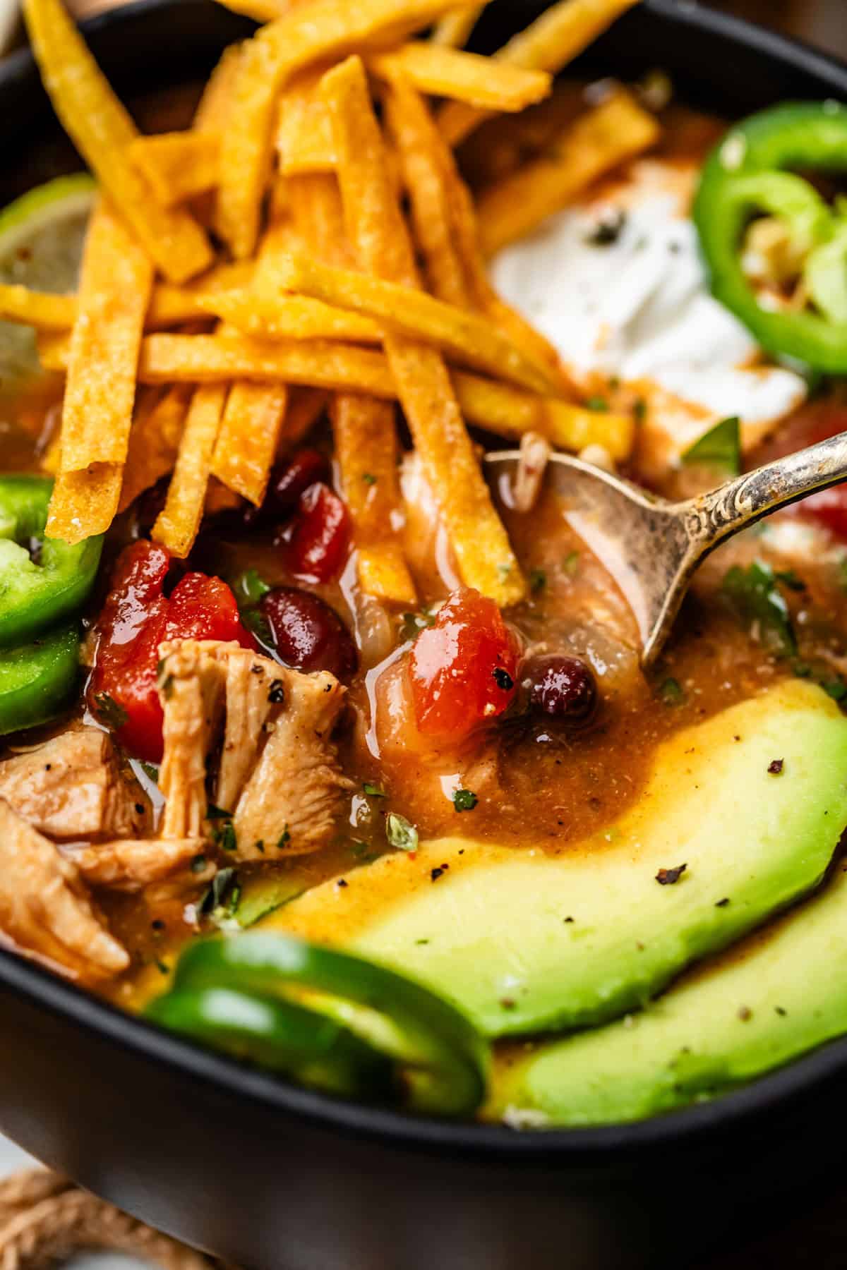close up of a metal spoon dipping into a bowl of chicken tortilla soup with toppings.