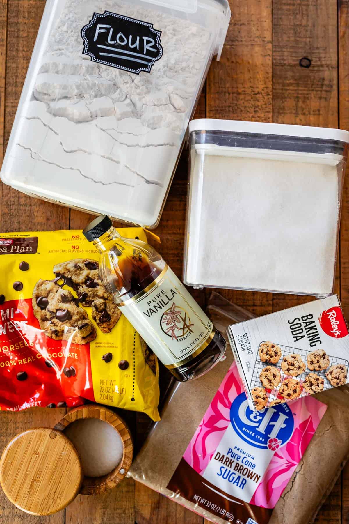 Ingredients for cookies laid out on a wood table: flour, sugar, brown sugar, baking soda, vanilla, chocolate chips, salt.