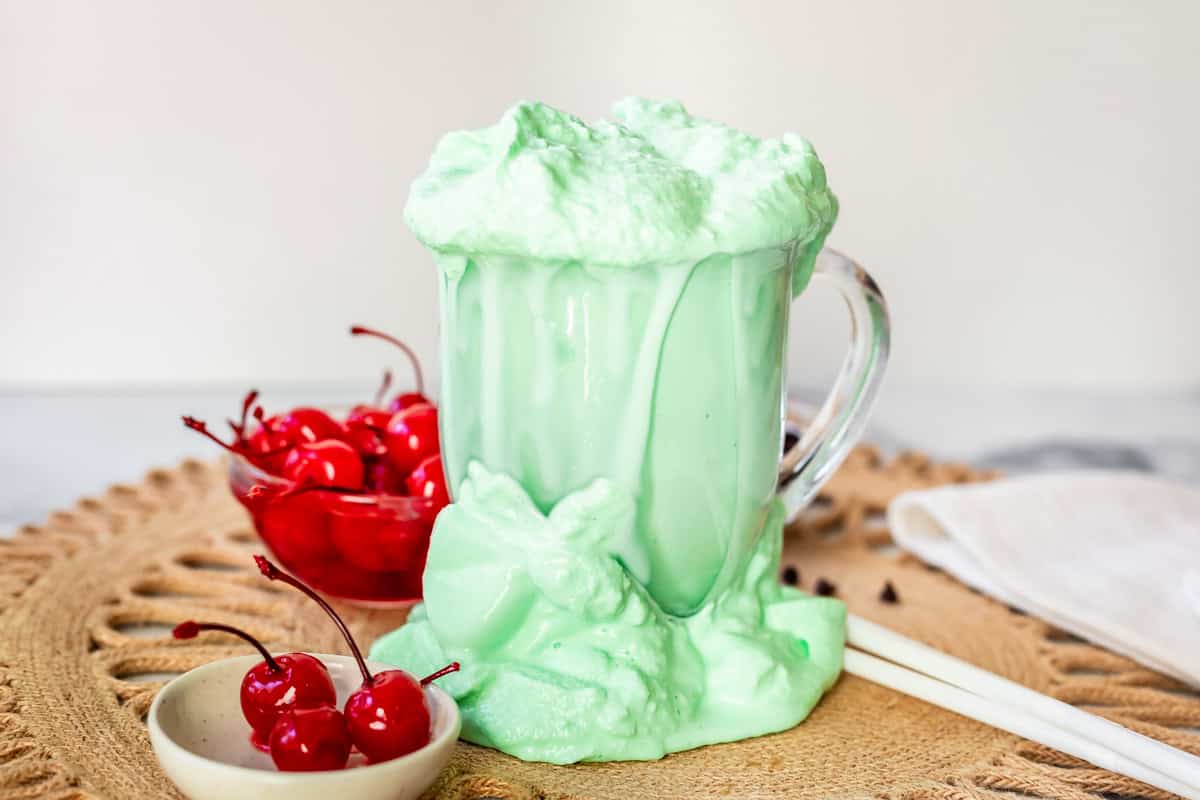shake overflowing and spilled all around a glass mug with bowl of cherries next to it.