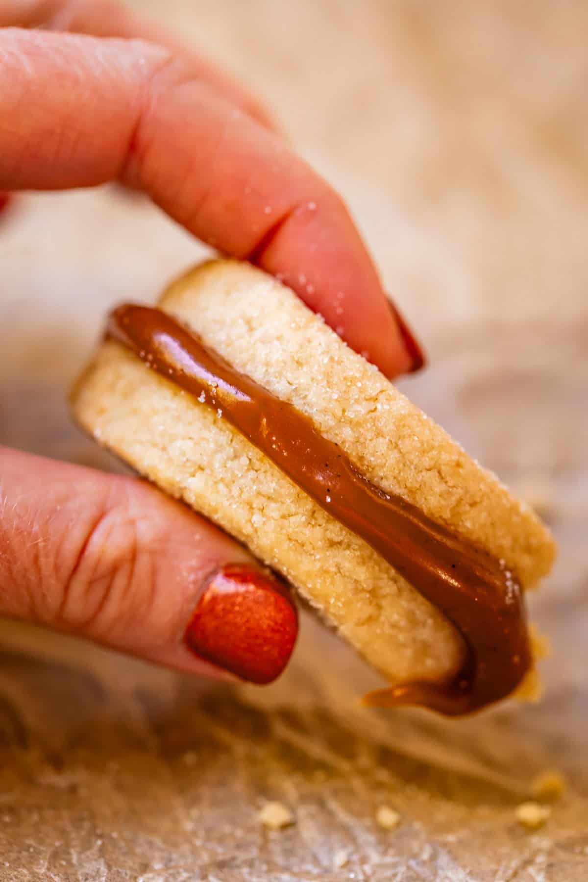 fingers holding up shortbread sandwich cookie with caramel in the center.