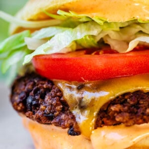 A close-up of a black bean burger with a crispy patty, melted cheese, creamy sauce, tomato slices, lettuce, and a soft bun. Some sauce is dripping down the side onto the plate.