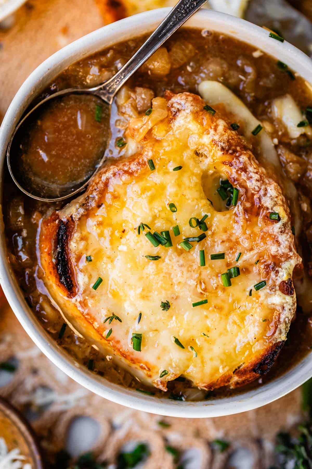 metal spoon holding french onion soup above bowl with bread and cheese.
