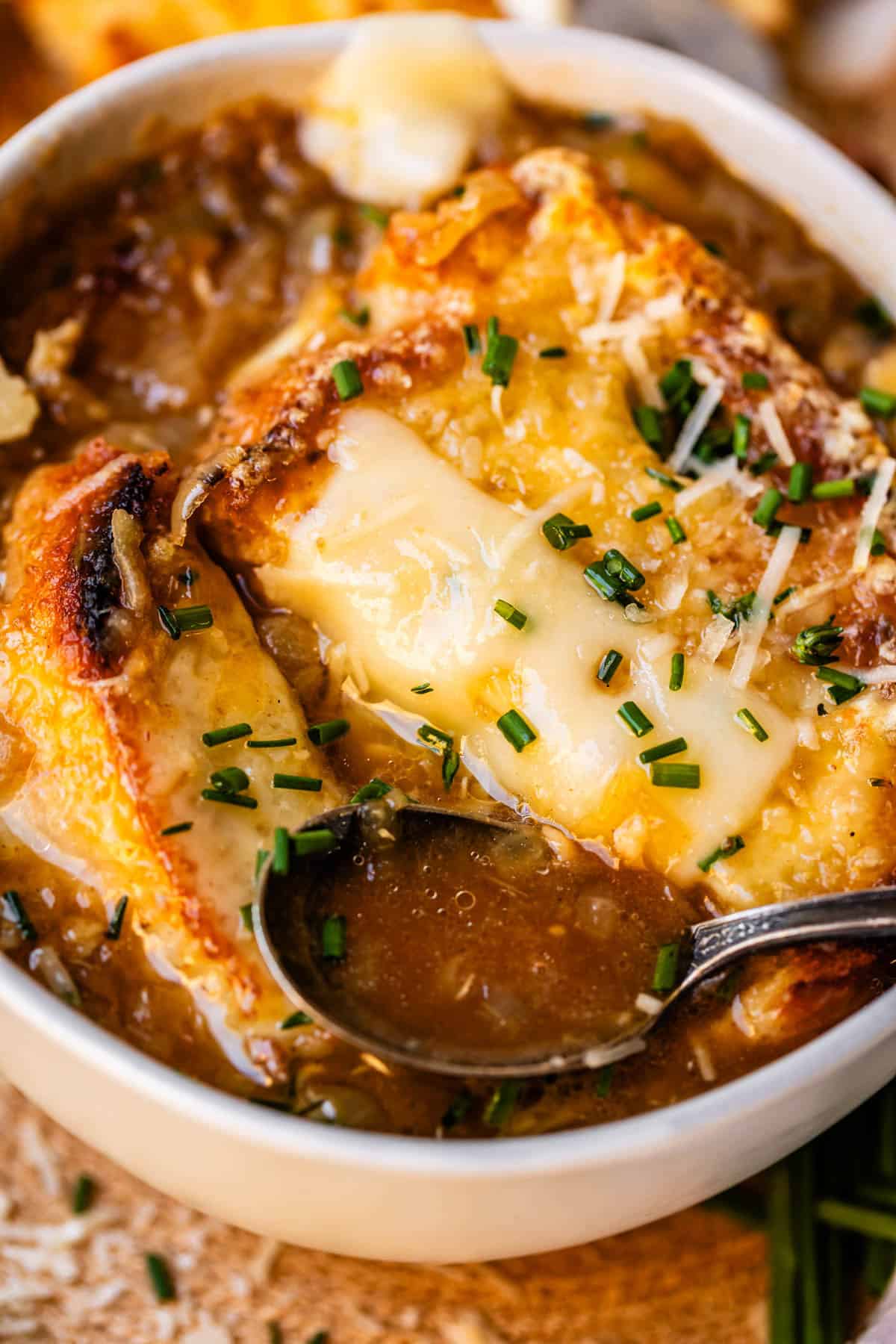 metal spoon holding french onion soup above bowl with bread and cheese.