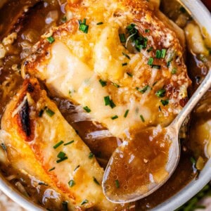 metal spoon holding french onion soup above bowl with toasty bread and cheese.
