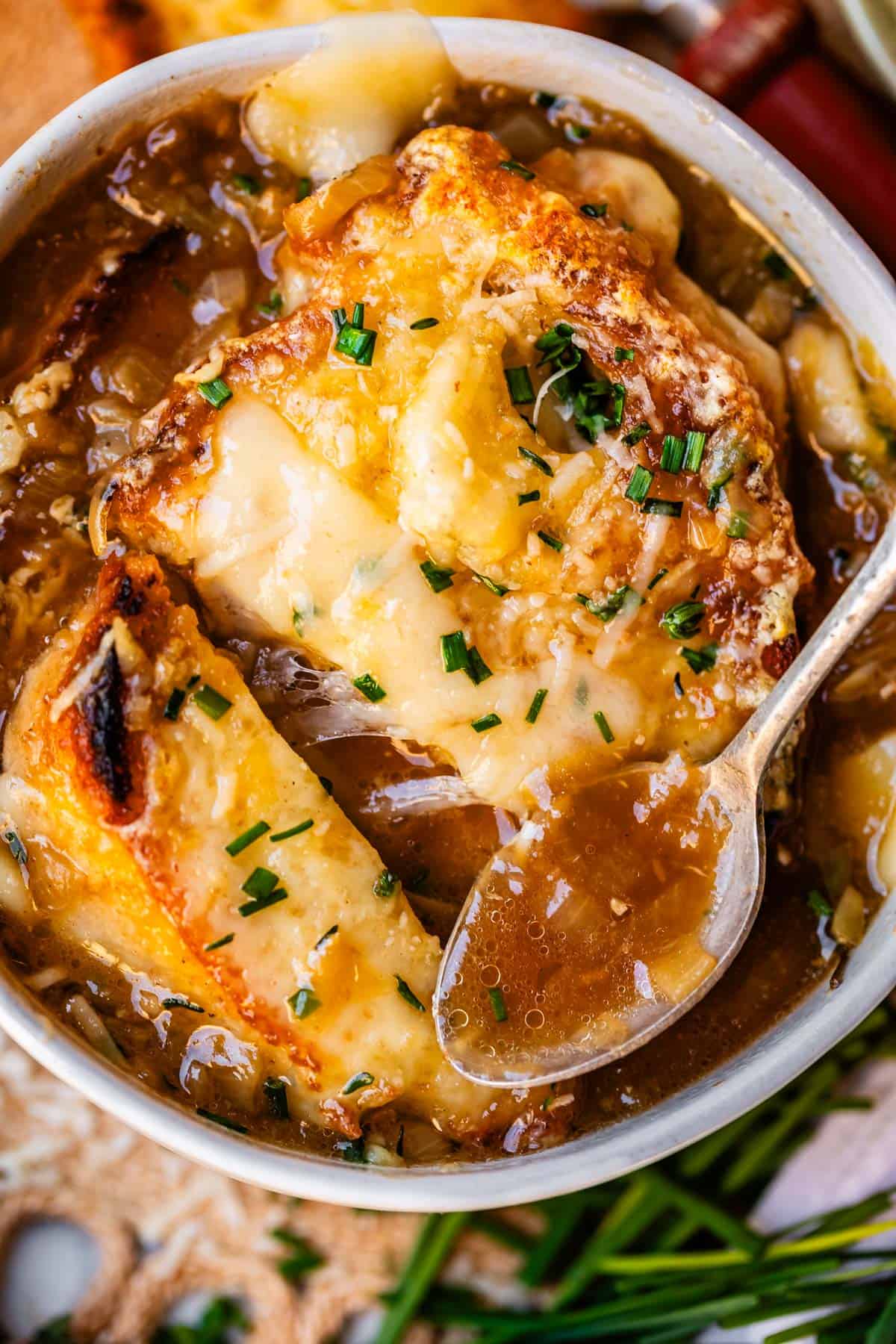 metal spoon holding french onion soup above bowl with toasty bread and cheese.