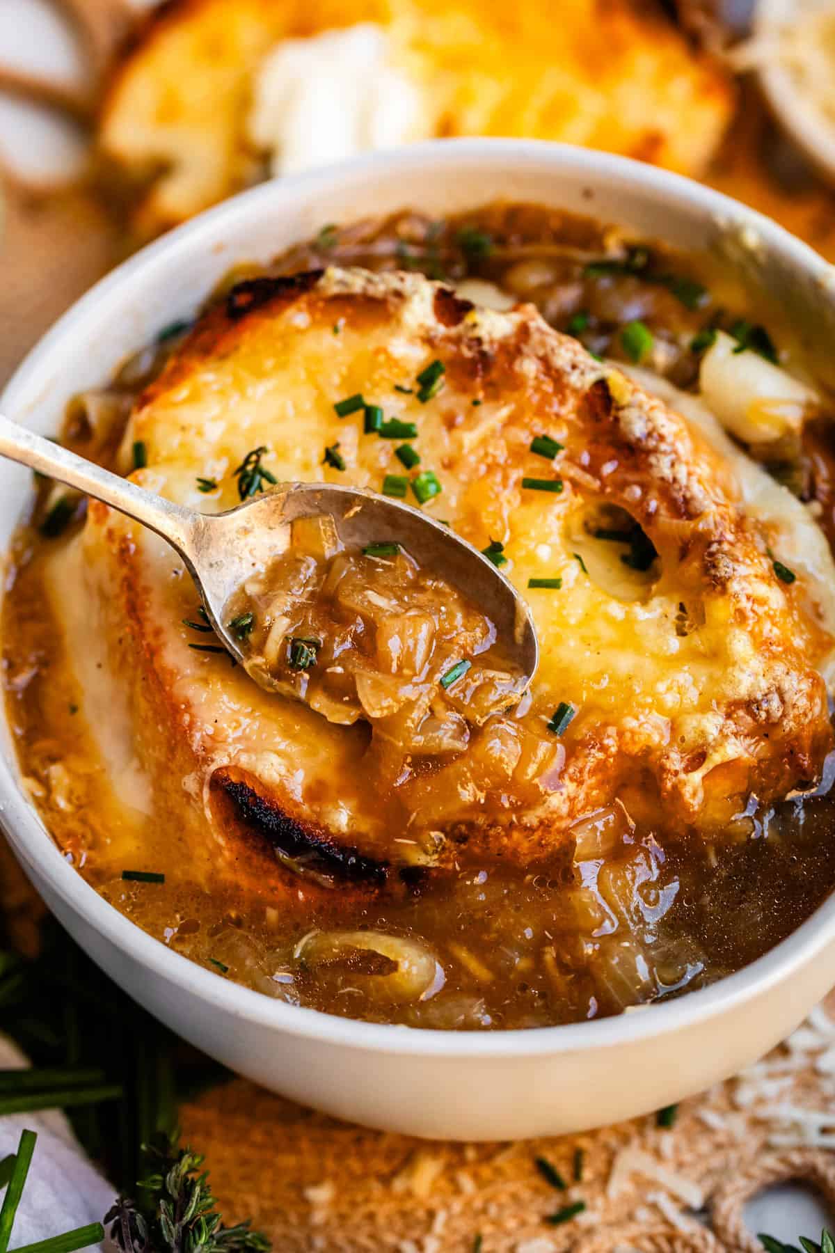 metal spoon holding french onion soup above bowl with toasty bread and cheese.