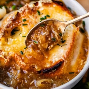 metal spoon holding french onion soup above bowl with toasty bread and cheese.