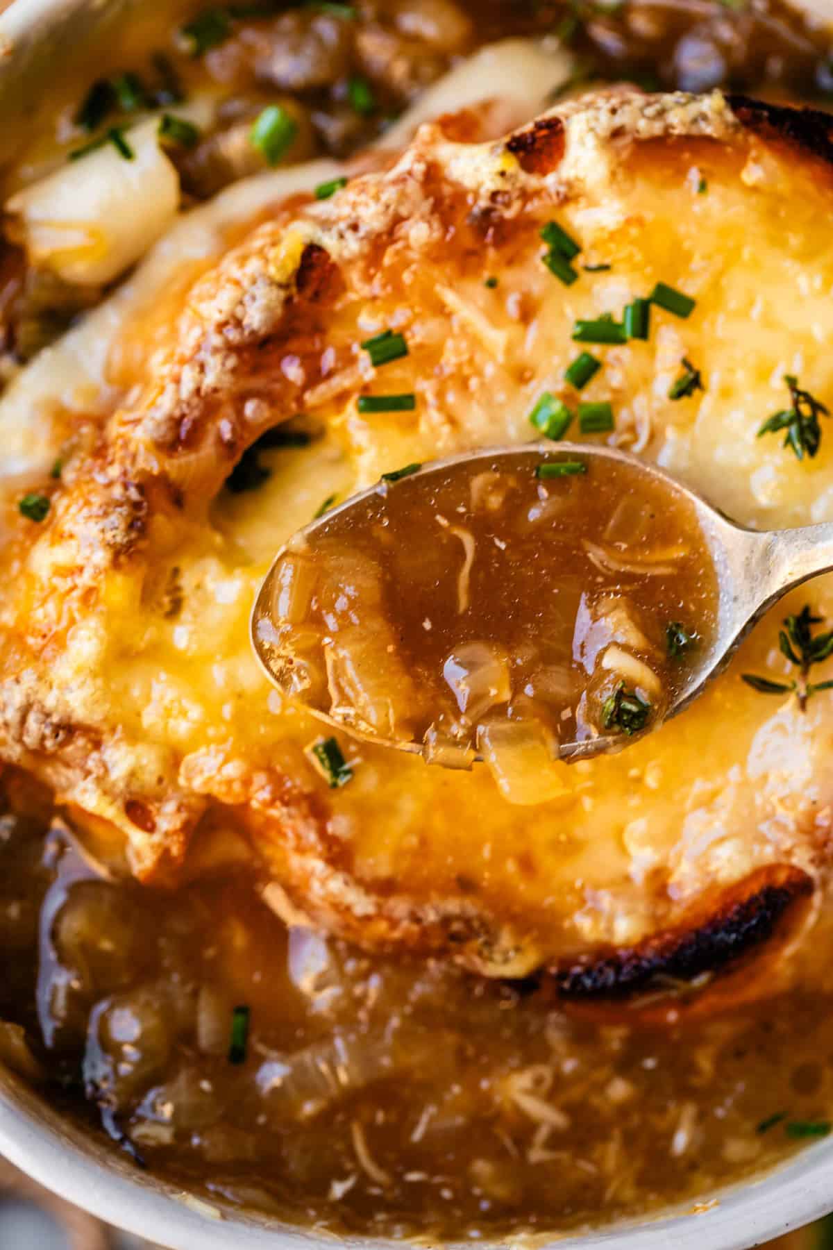 metal spoon holding french onion soup above bowl with toasty bread and cheese.
