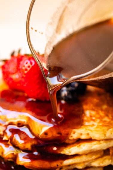 Homemade maple syrup being poured out of a glass spout over pancakes with strawberries in the background.