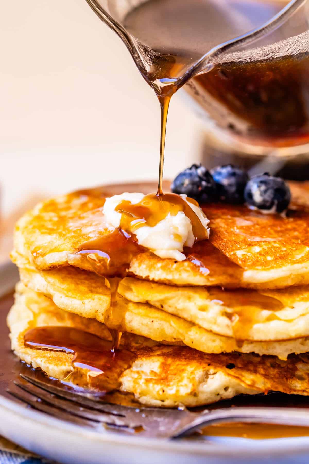 pouring maple syrup over pancakes with butter and blueberries.