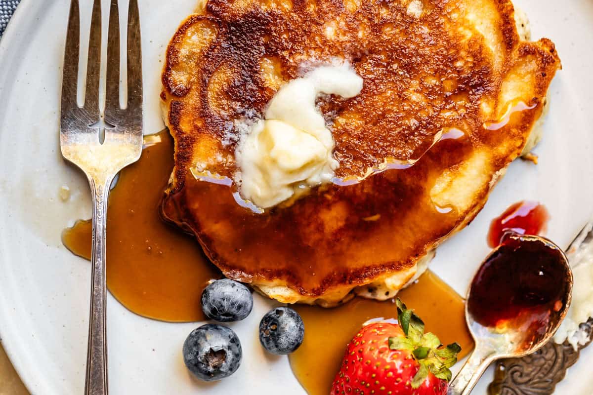Browned pancake on a plate with butter, syrup, fork & knife, and fruit.