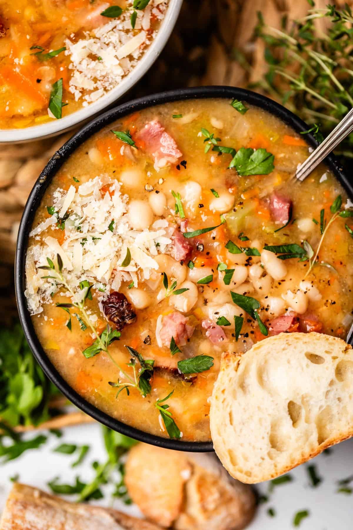 ceramic bowl of ham & bean soup garnished with cheese, pepper, and a slice of french bread.