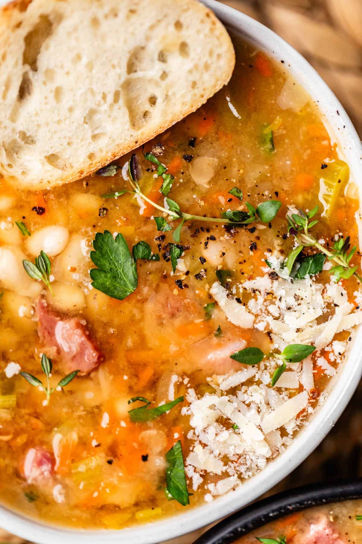 close up of cheese and pepper garnished ham & bean soup with a piece of bread on side of bowl.