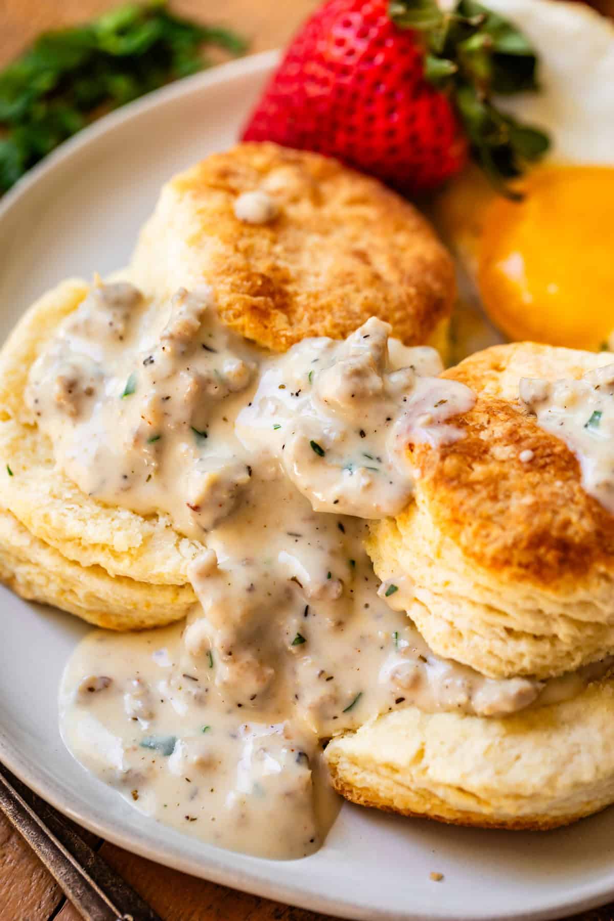 two biscuits, gravy, and strawberries on a ceramic dinner plate on a wood table next to a fork.