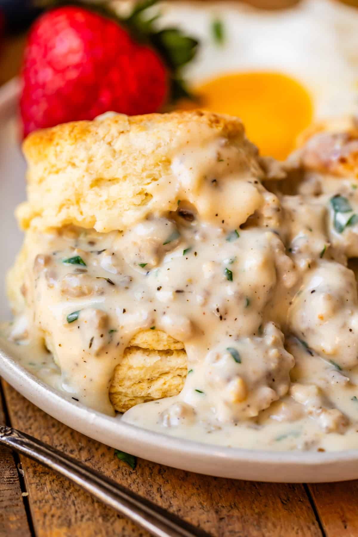Biscuits, gravy, and strawberries on a ceramic dinner plate on a wood table next to a fork.