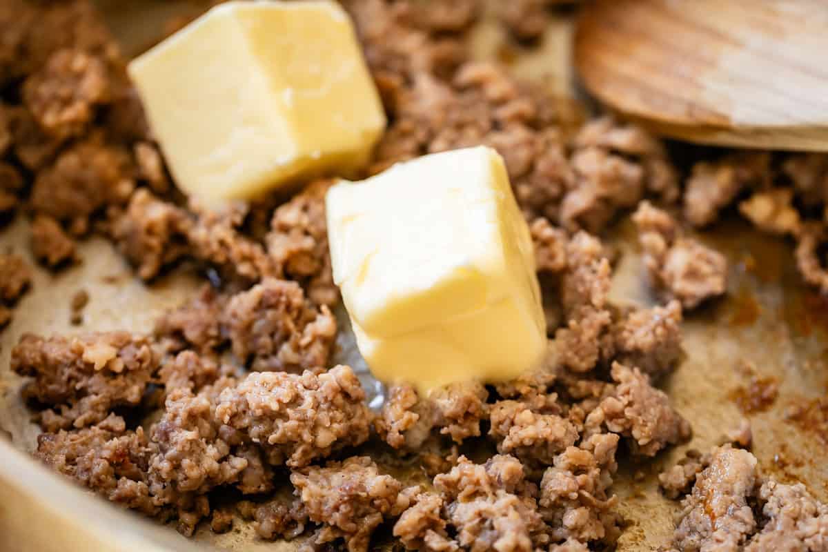 two large pats of butter melting into cooked sausage in a metal skillet.