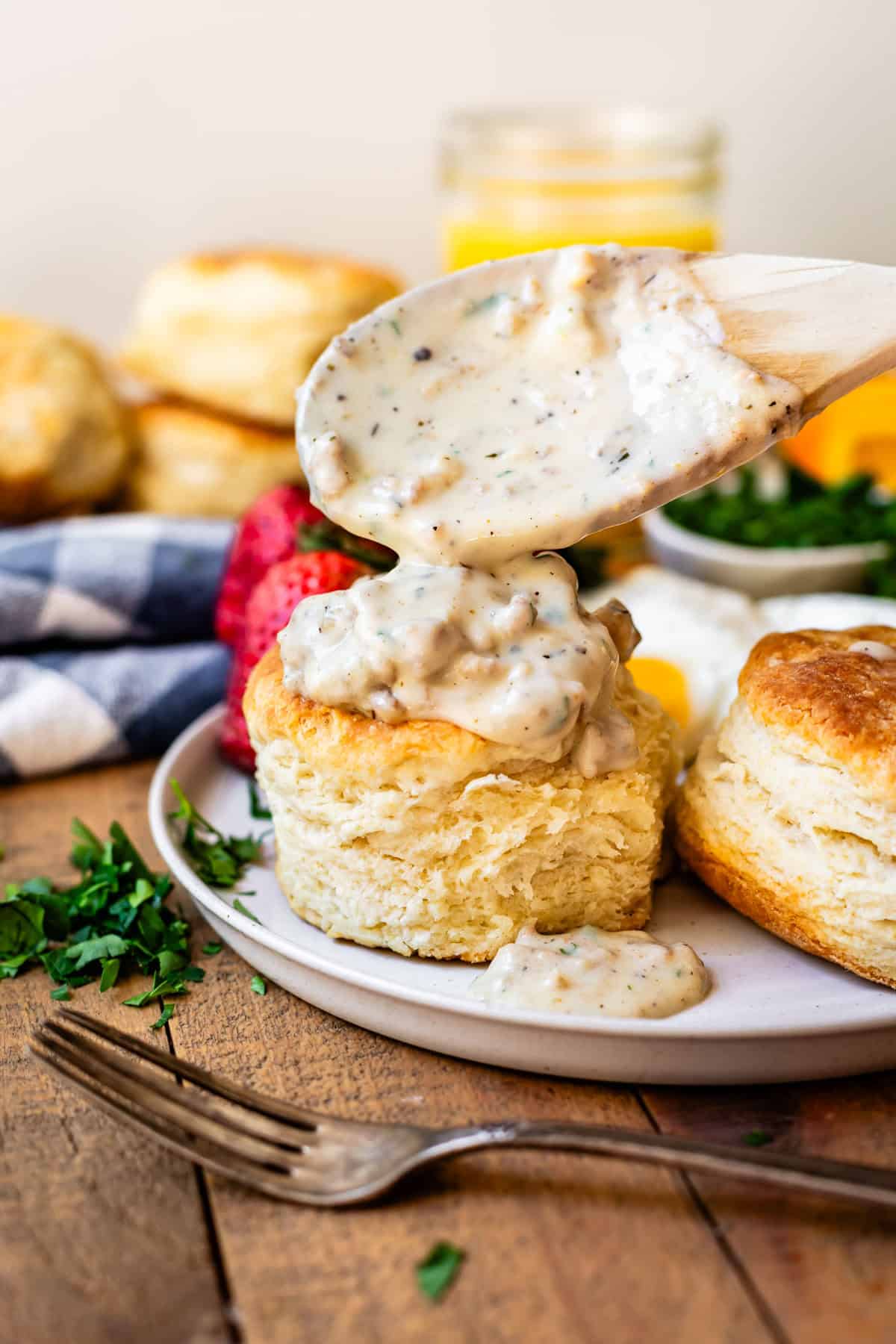 wooden spoon pouring sausage gravy over the top of biscuits on a ceramic plate.