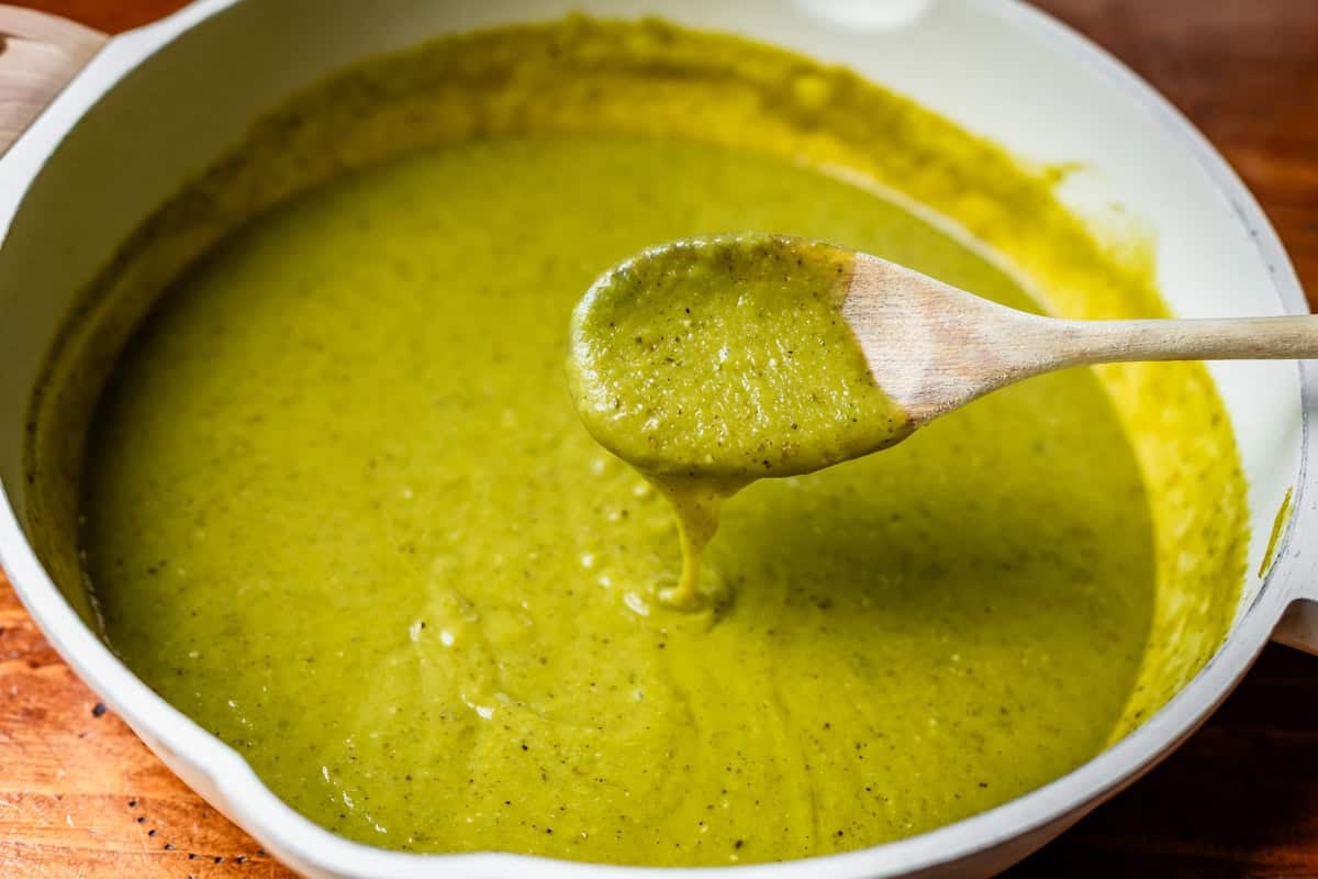 wooden spoon dipping into blended green enchilada sauce in a frying pan.