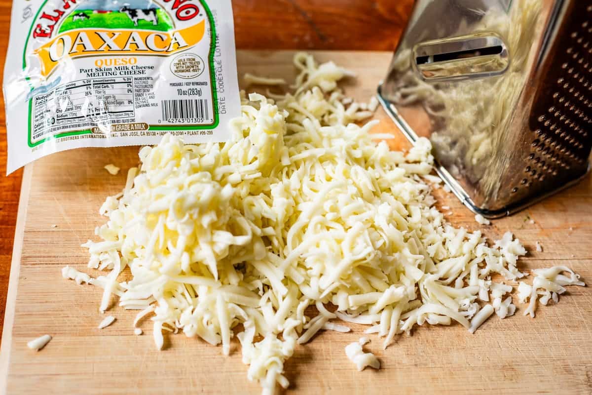 shredded Oaxaca cheese on a wood cutting board next to a box cheese grater.