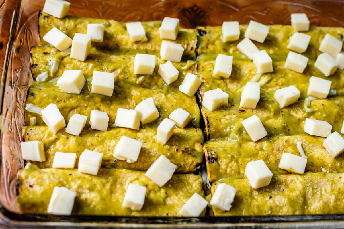 cubed monterey jack cheese spread on top of unbaked green chicken enchiladas in glass pan.