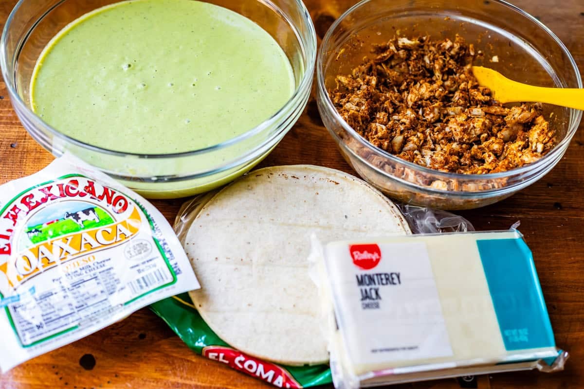 bowl of green enchilada sauce, bowl of seasoned chicken, tortillas, and cheese on a table.