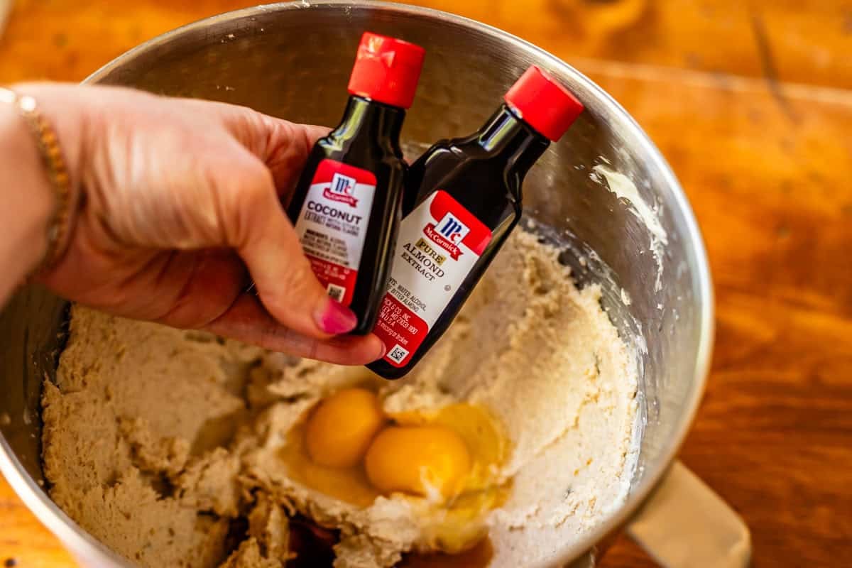 hand holding coconut and almond extract bottles over the mixing bowl with dough in it.