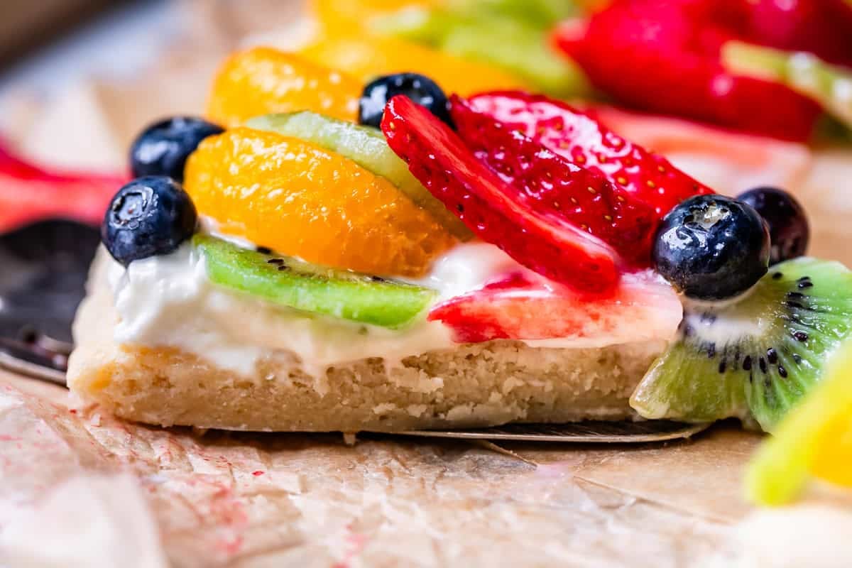 close up of a side view of fruit pizza showing the cookie base, frosting, and layers of fruit.