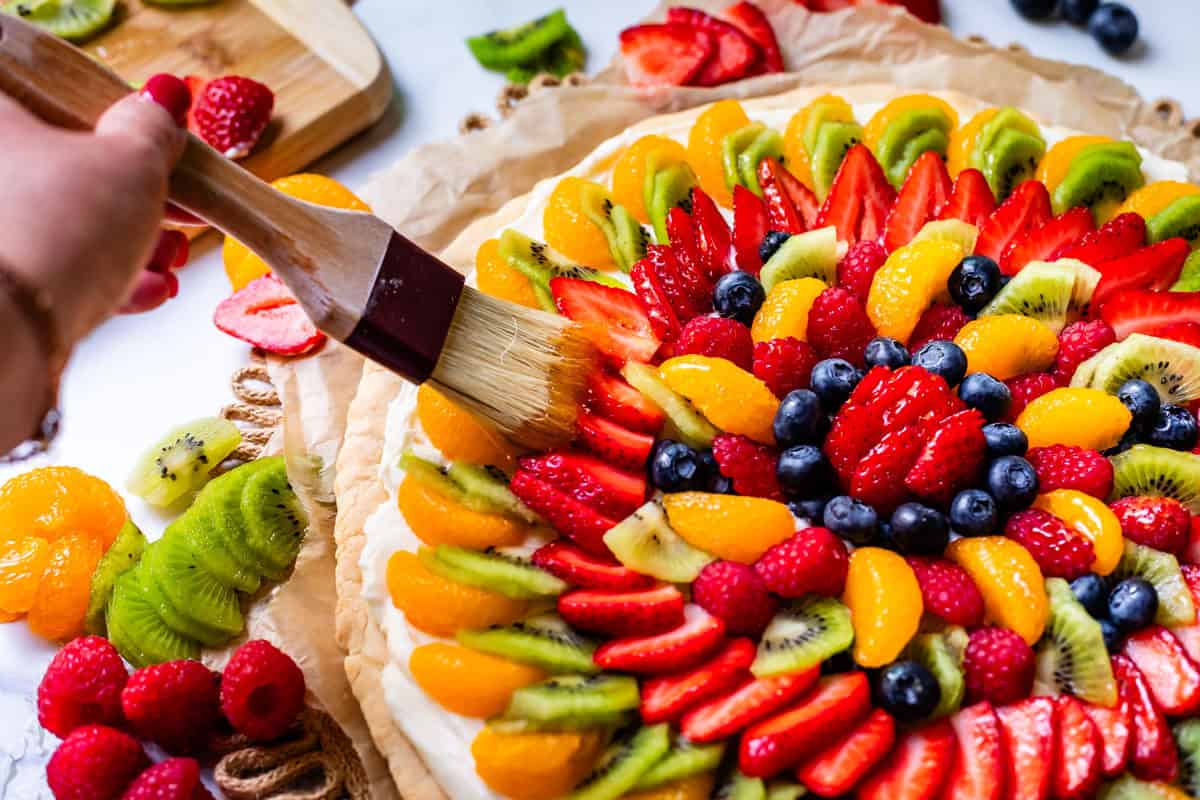hand holding pastry brush to brush glaze on the fruit on top of the baked and frosted cookie crust.